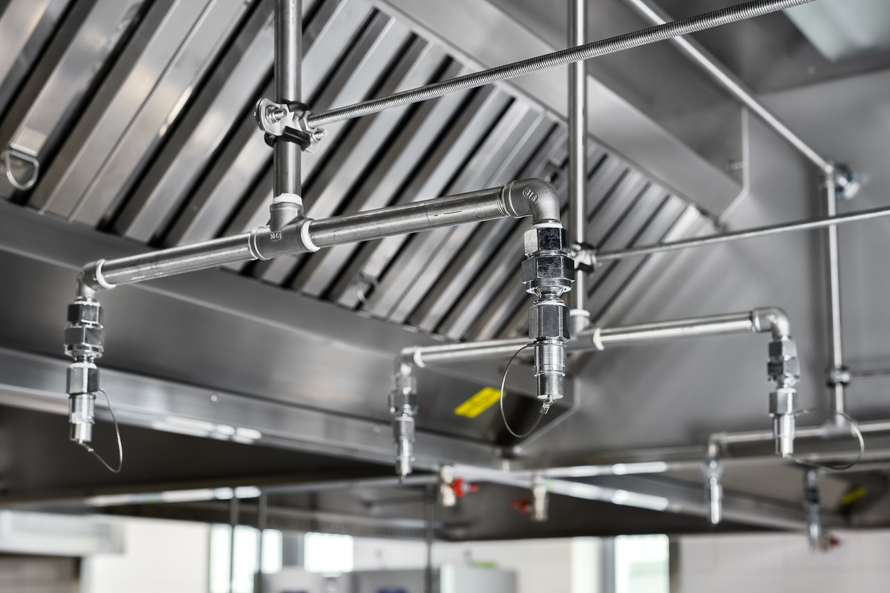 Close-up of stainless-steel kitchen ventilation hood with fire-suppression piping and nozzles — training kitchen by GORT